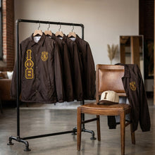 Load image into Gallery viewer, Brown jackets with yellow logos on a rack and chair in a room.