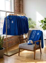 Load image into Gallery viewer, Blue shirts with a logo on a rack and chair in a room with plants and a brick wall.
